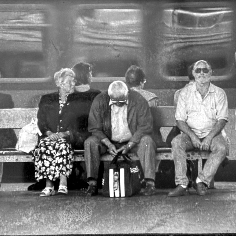 Black-and-white photograph from 1999: people waiting on a platform bench, a passing train behind them